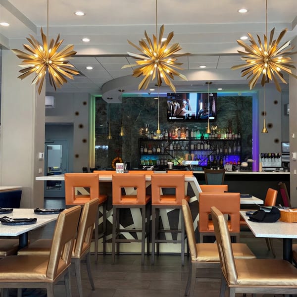 A modern restaurant interior with a bar, orange bar stools, and golden starburst chandeliers.