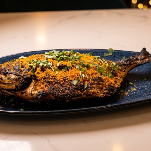 Grilled whole fish, seasoned with orange spices and green herbs, served on a dark blue plate.