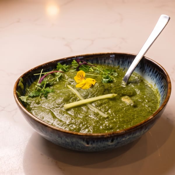 A bowl of vibrant green curry, garnished with herbs, a yellow edible flower, and green stalks.
